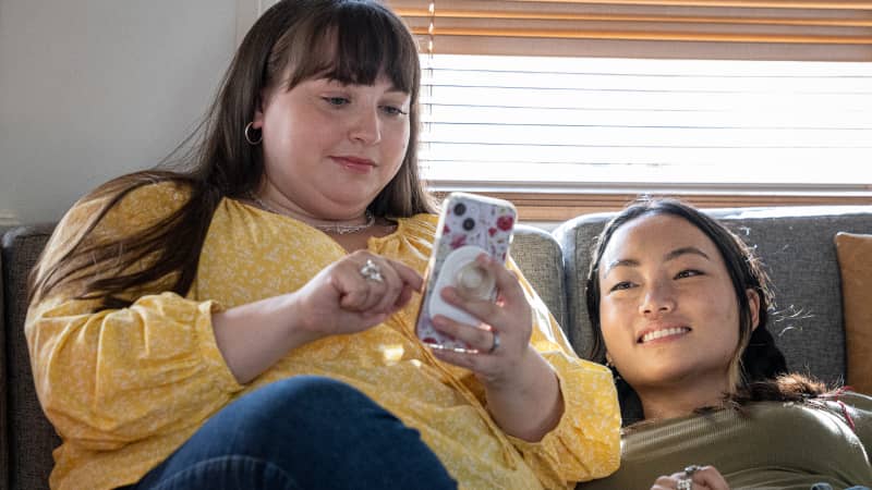 Two young people on a sofa, checking order status on the screen of a cell phone one is holding.