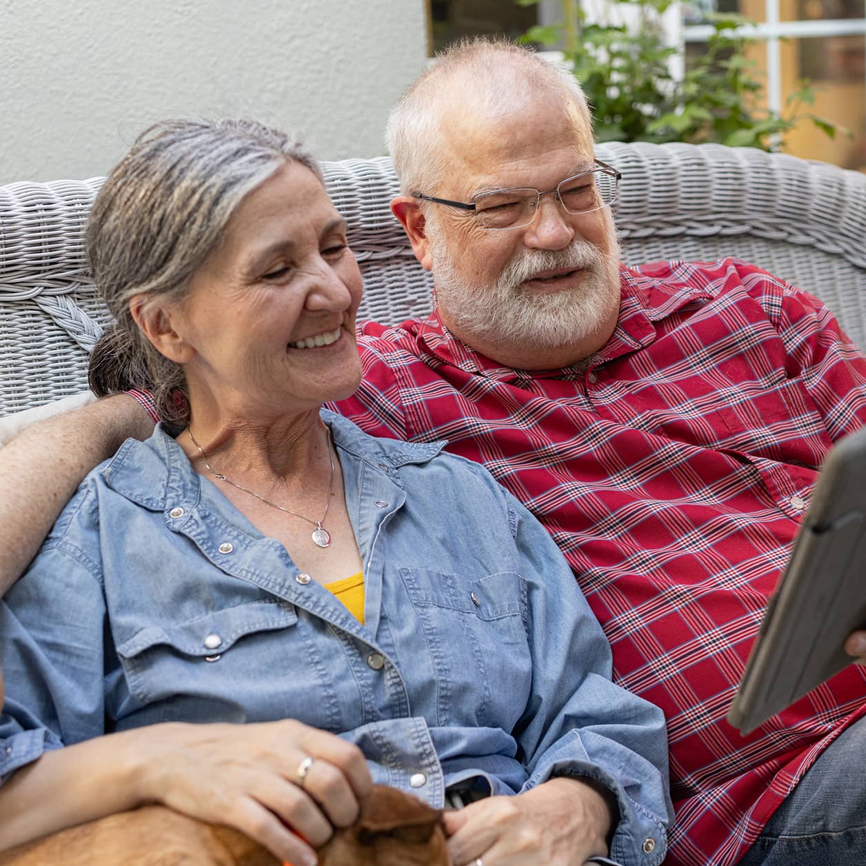 An older smiling couple on a sofa, looking at a tablet screen.