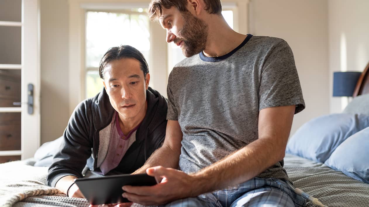 A couple looks at a tablet screen together sitting on their bed.
