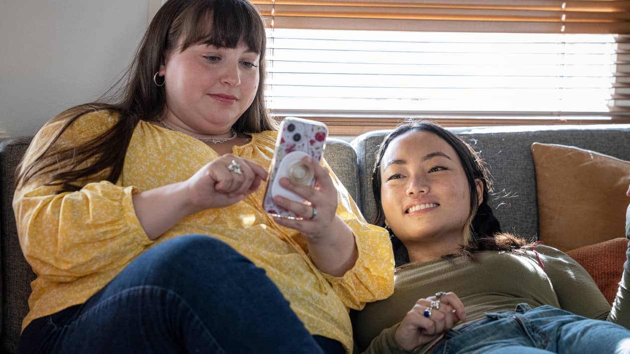 Two young people on a sofa, checking order status on the screen of a cell phone one is holding.