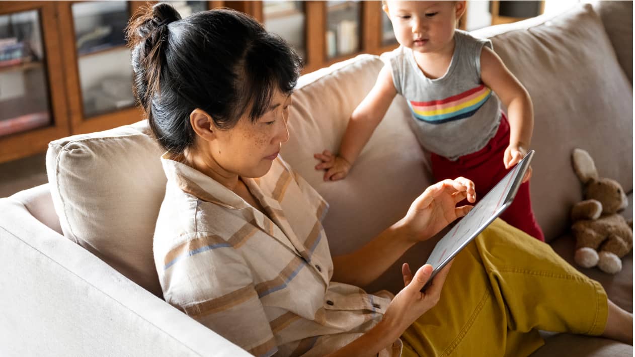 A parent on the sofa with their toddler, and looking at a tablet screen.