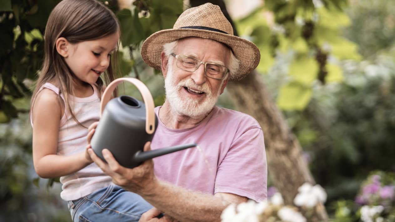 A grandparent holds a grandchild as they water plants.