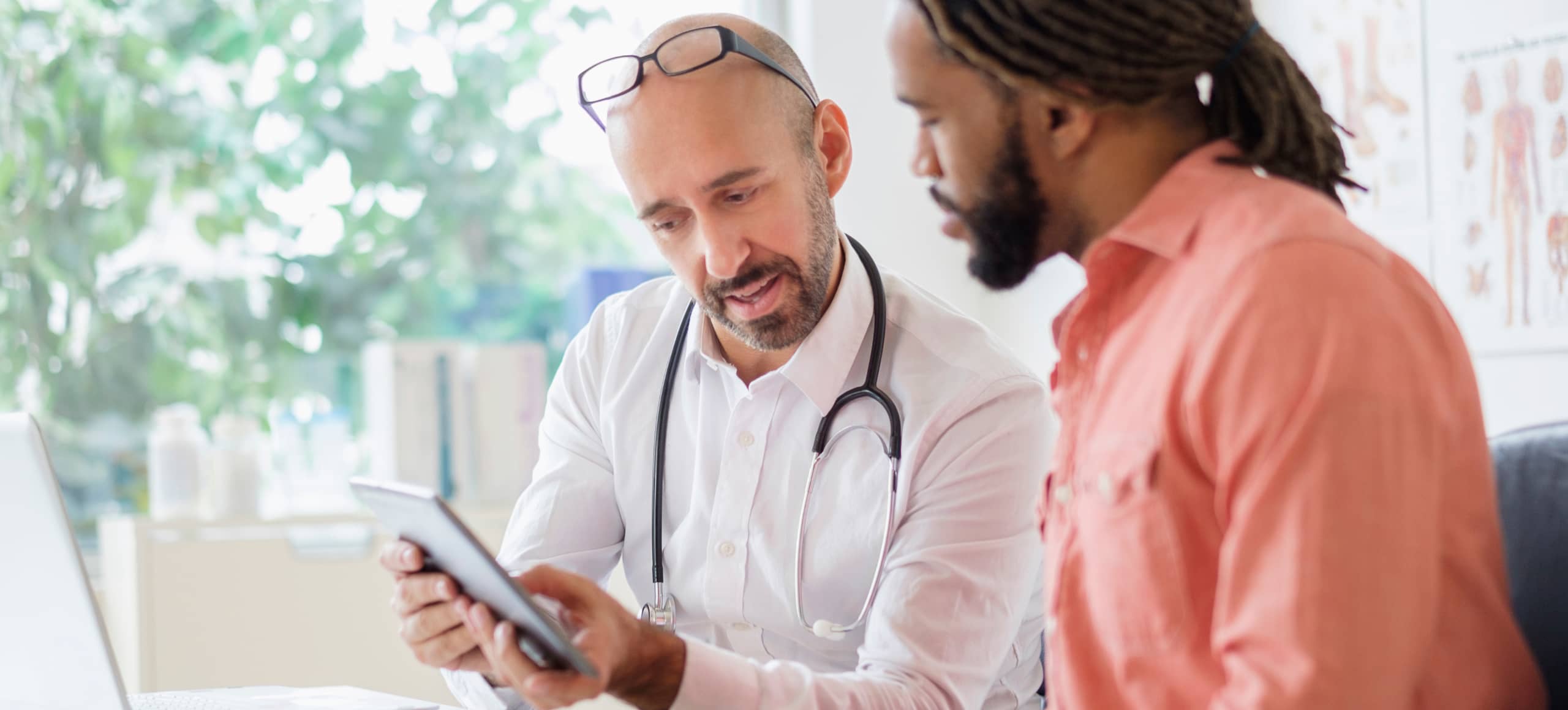A practitioner wearing a stethoscope, showing a tablet screen to the patient beside them.