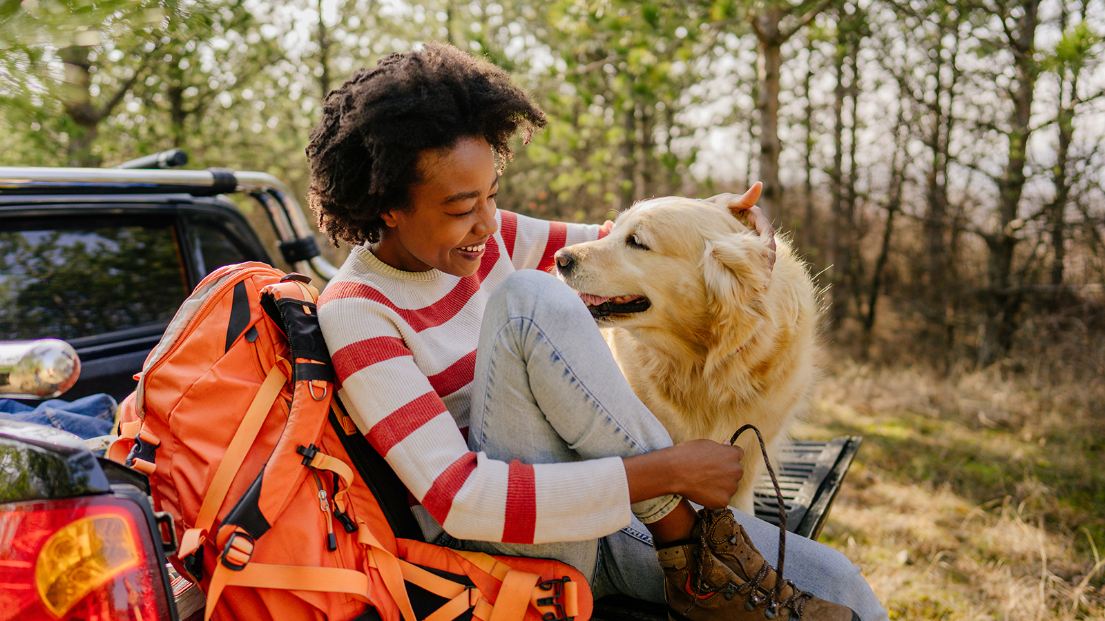 A person sits on a truck bed with an orange backpack, petting a golden retriever in a forested area.