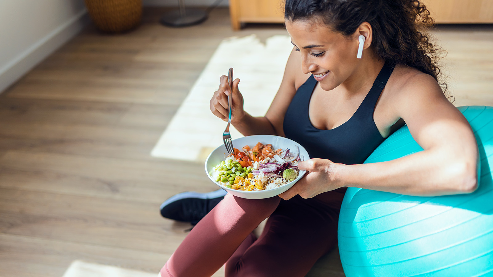 A woman in athletic wear sits on the floor, eating a colorful bowl of food while resting against a blue exercise ball.