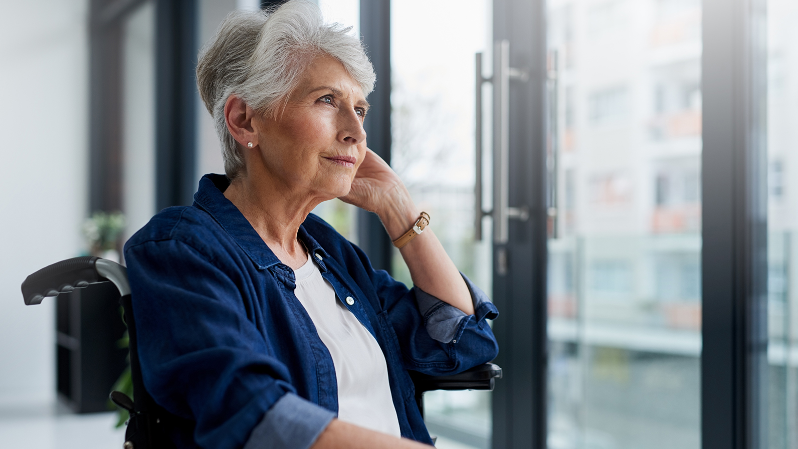 An elderly woman sits in a wheelchair, looking thoughtfully out of a large window with natural light streaming in.