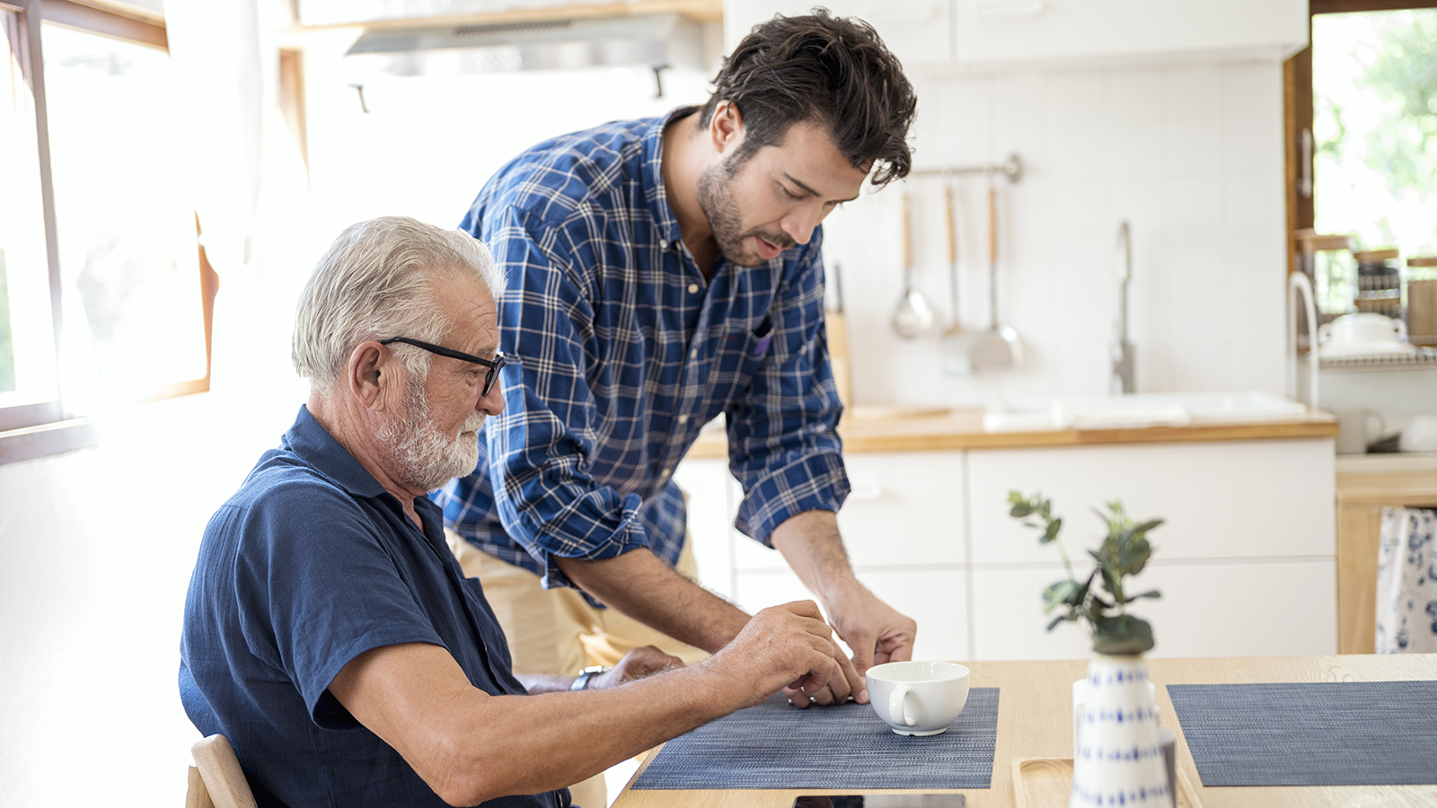A younger man assists an older man at a kitchen table, placing a cup in front of him.