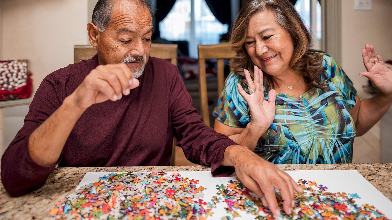 Two people are seated at a table, gesturing excitedly over a colorful jigsaw puzzle spread out before them.