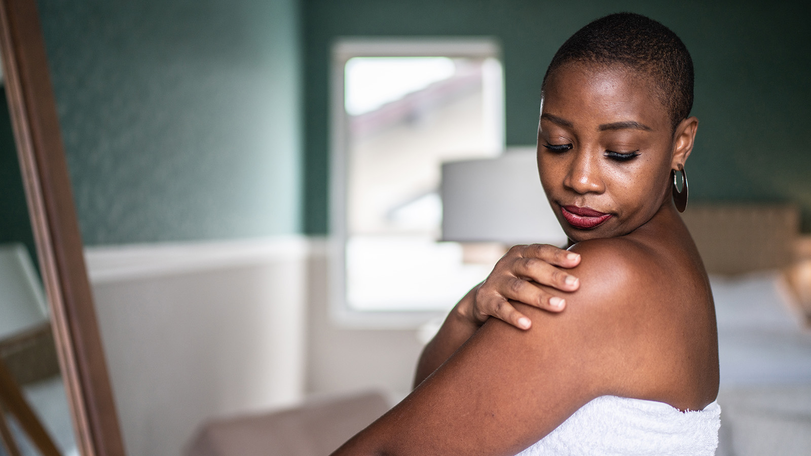 A person stands in front of a mirror, gently touching their shoulder, with a soft-focus background of a room.