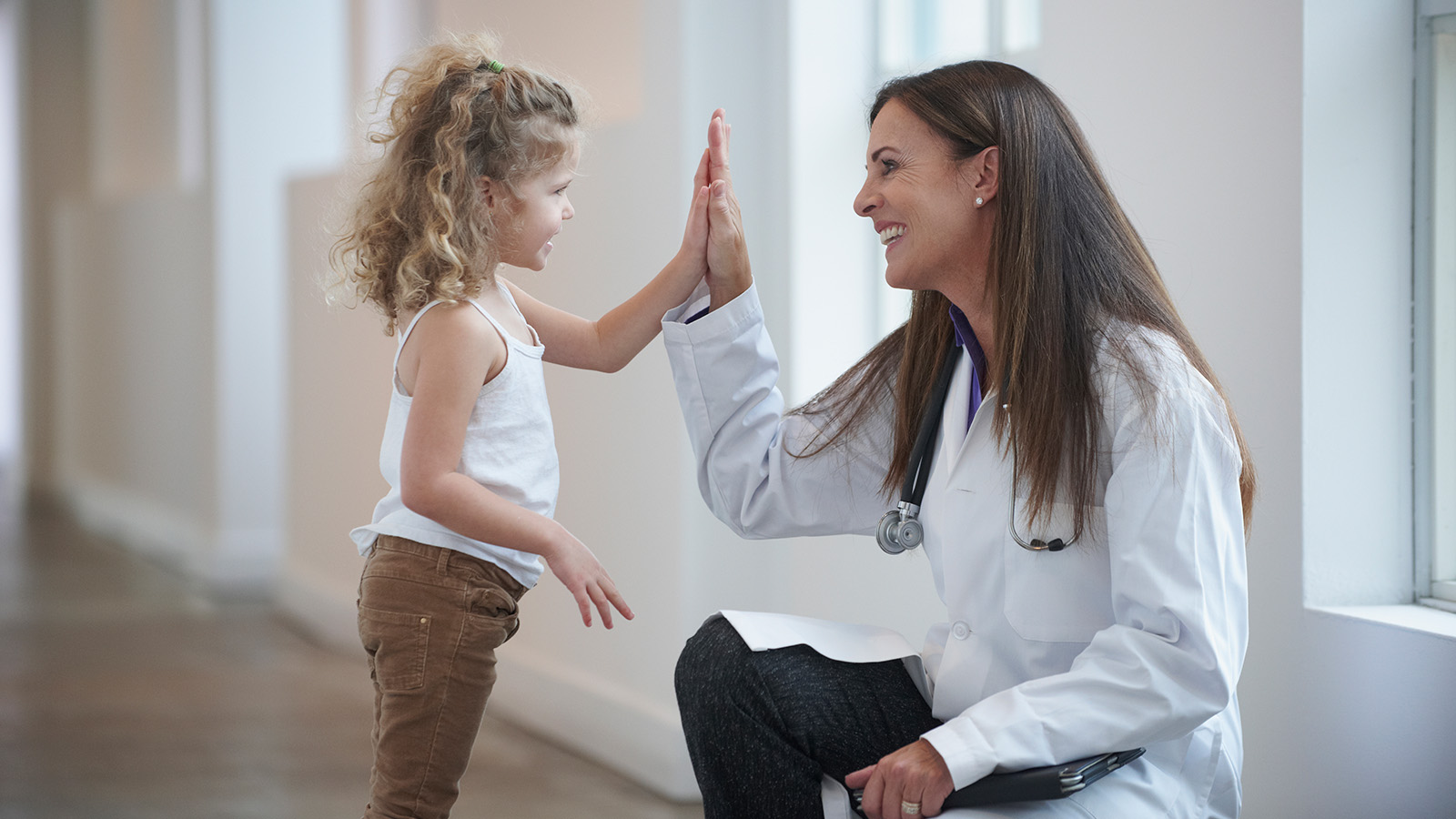 A doctor and a young girl are giving each other high fives in a bright hallway.