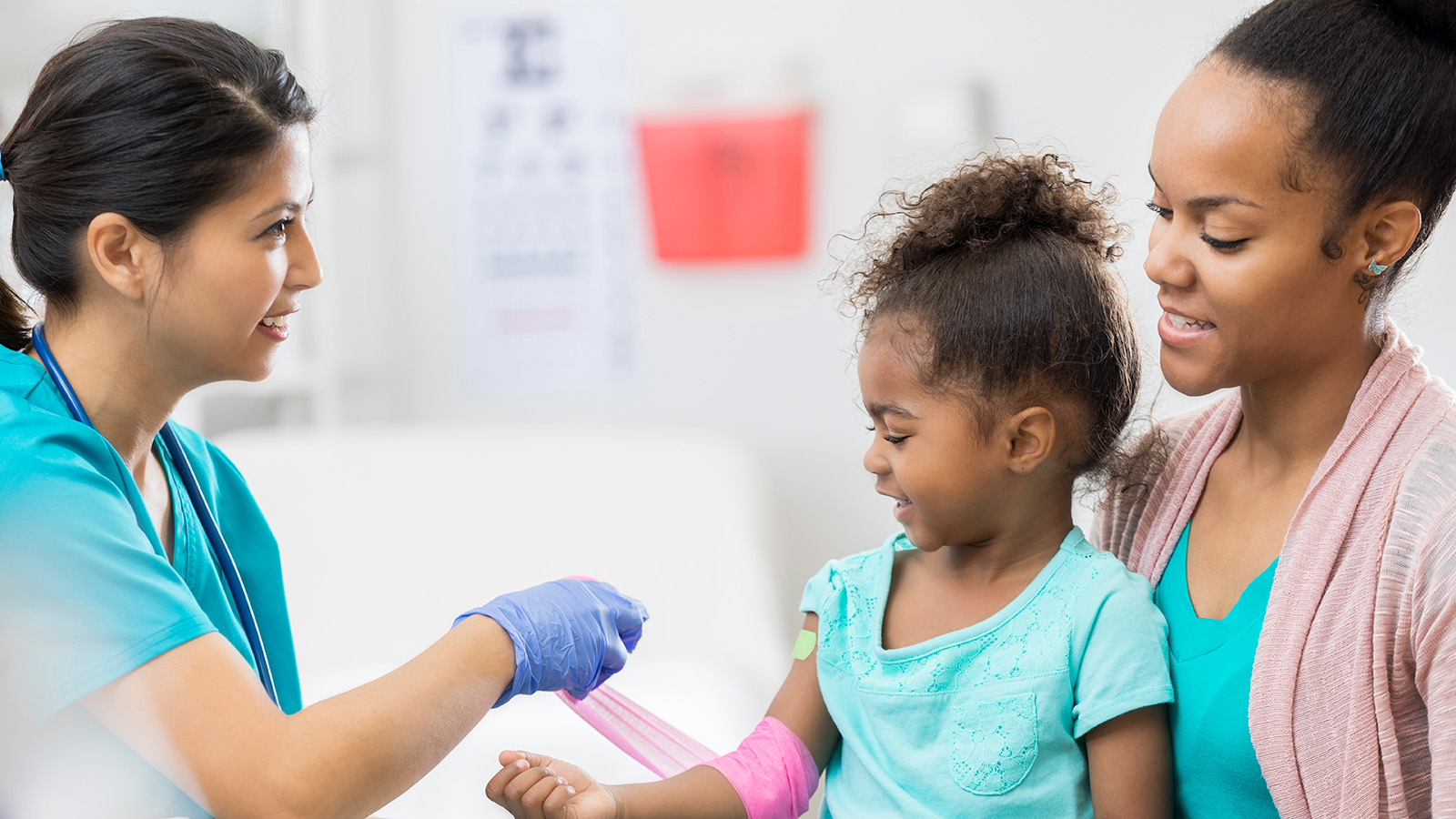 A nurse applies a bandage to a child’s arm while the child’s mother watches closely in a medical setting.