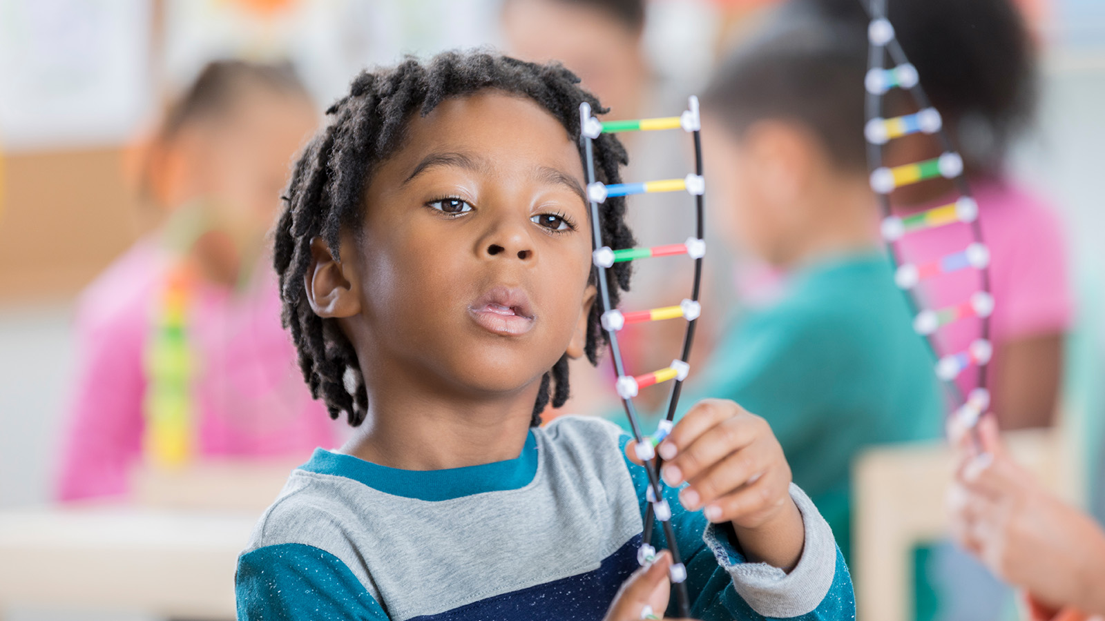 A child holds a colorful model of DNA while other children are engaged in activities in the background.
