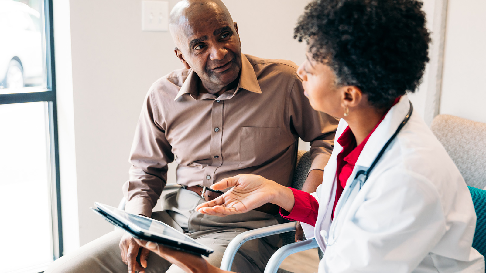 A doctor discusses health with a patient in a waiting room, holding a tablet and gesturing with her hand.