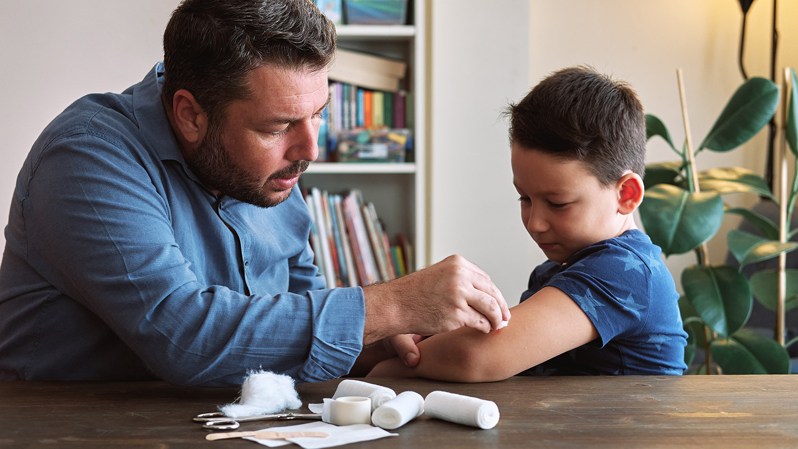 An adult applies a bandage to a child’s arm, with medical supplies laid out on a table nearby