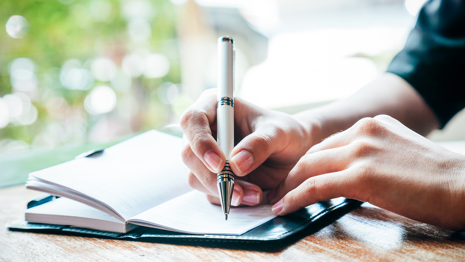 A person writes in a notebook with a pen, focusing intently on the task, with a blurred background.