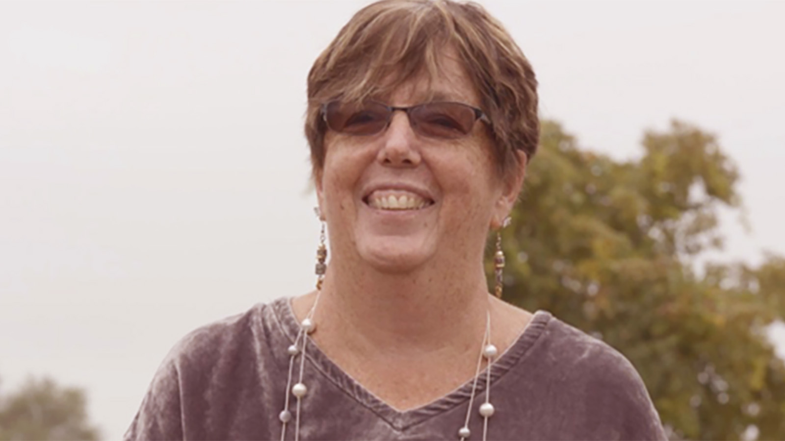 A woman stands outdoors, wearing a gray top and long earrings, with trees and a blurred background of nature.