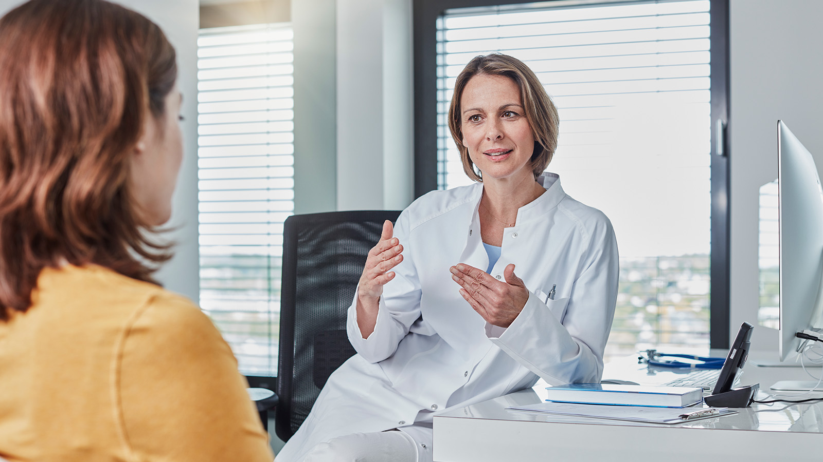 A doctor in a white coat gestures while explaining something to a patient sitting across from her in an office.