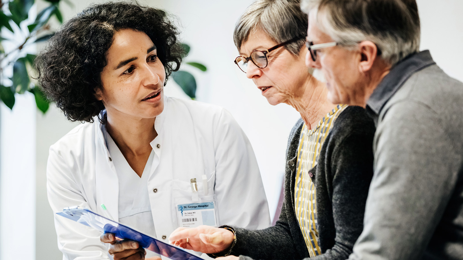 A doctor discusses information with an elderly couple, showing them a clipboard in a well-lit medical office.