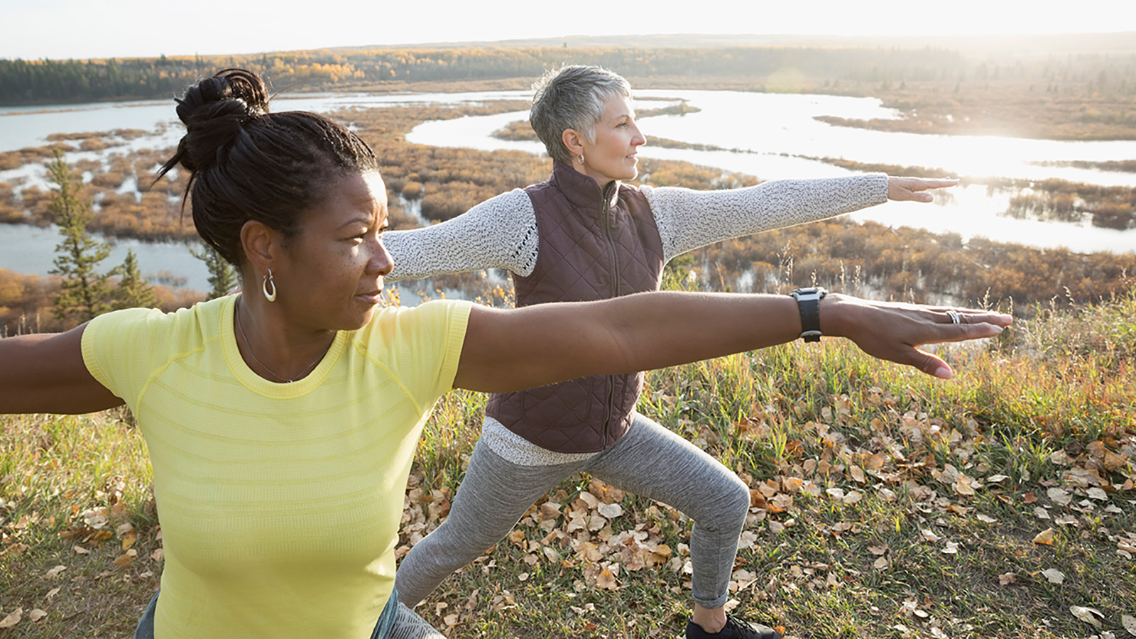 Two women practice yoga poses outdoors, stretching their arms in a scenic area with a river and autumn foliage.