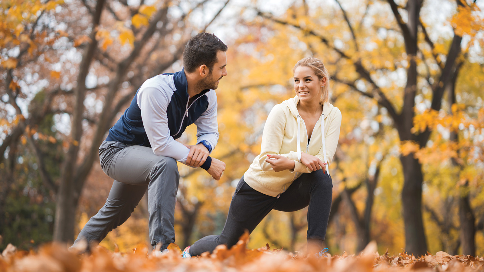 A man and woman stretch together outdoors in a park filled with autumn leaves, focusing on their fitness routine.