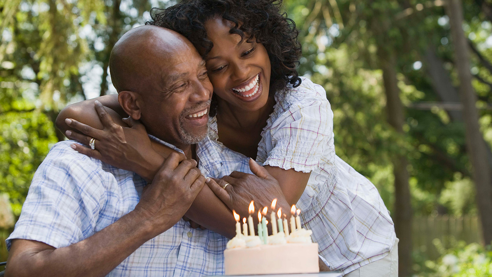 A couple embraces joyfully beside a birthday cake with lit candles outdoors, surrounded by greenery.