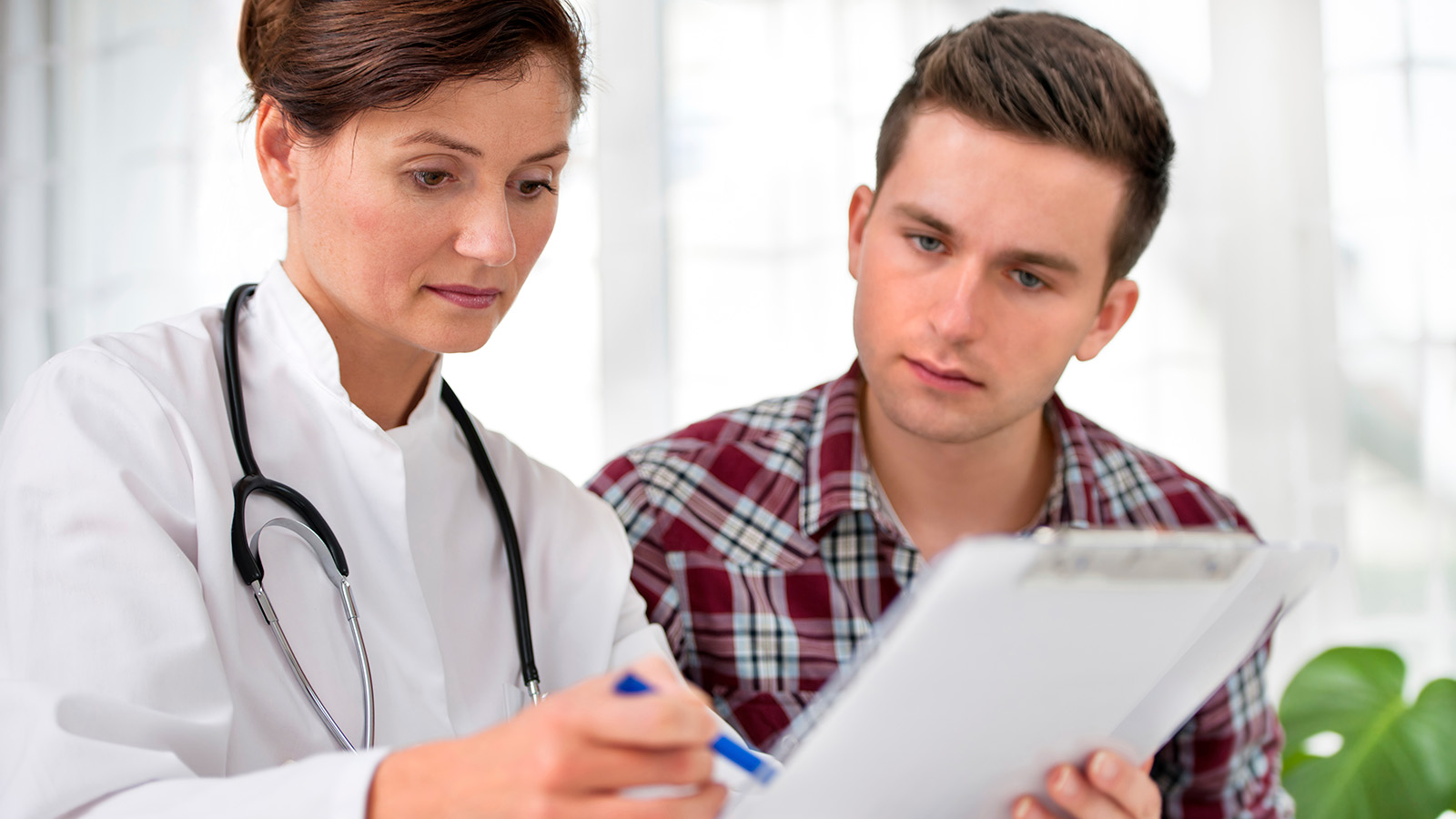 A doctor discusses medical information with a young man, both focused on a clipboard in a well-lit room.