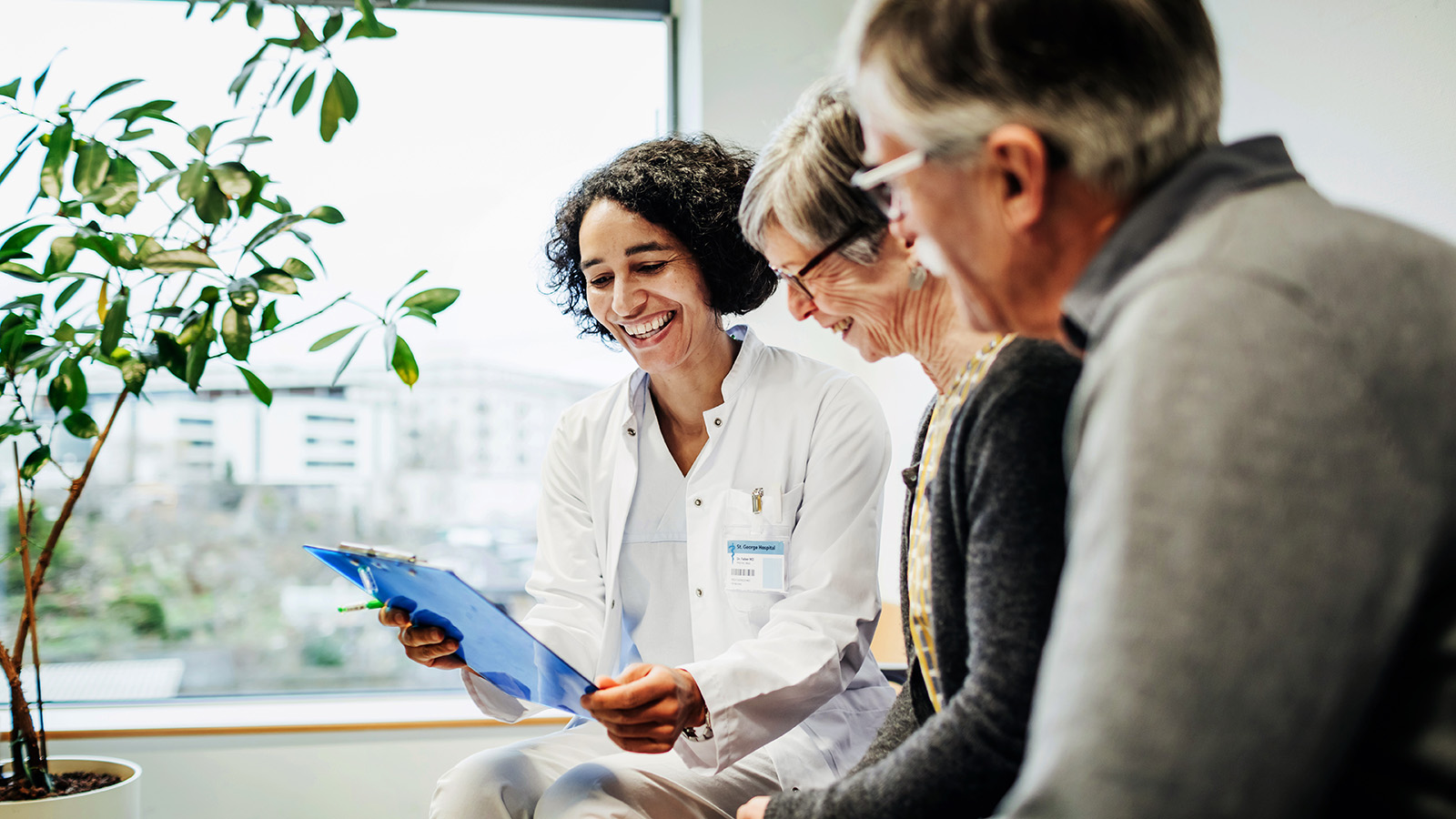 A medical professional explains information to an elderly couple, seated together in a comfortable waiting area.