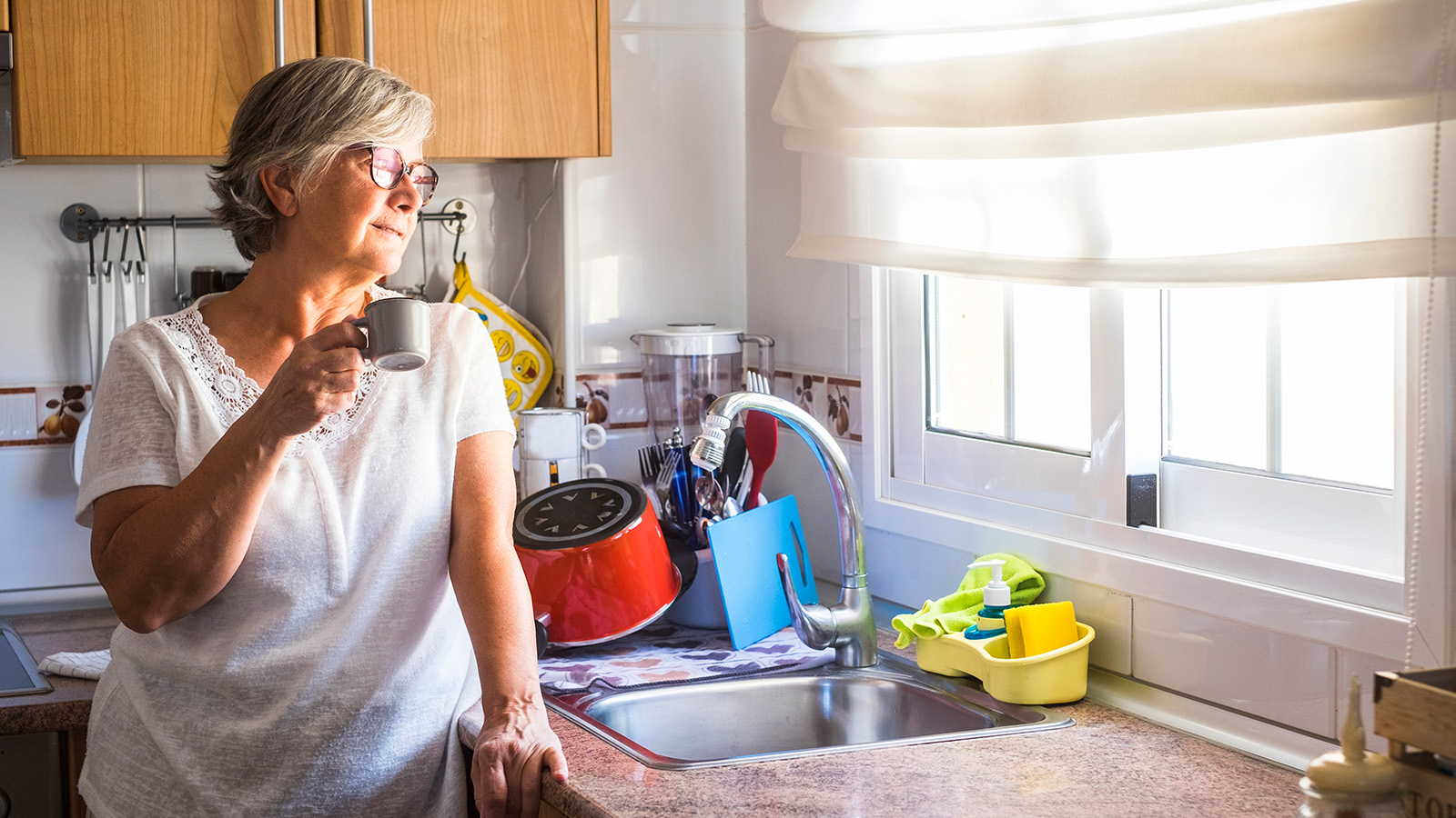 An older woman stands in a kitchen, sipping coffee while looking out the window, enjoying a peaceful moment.