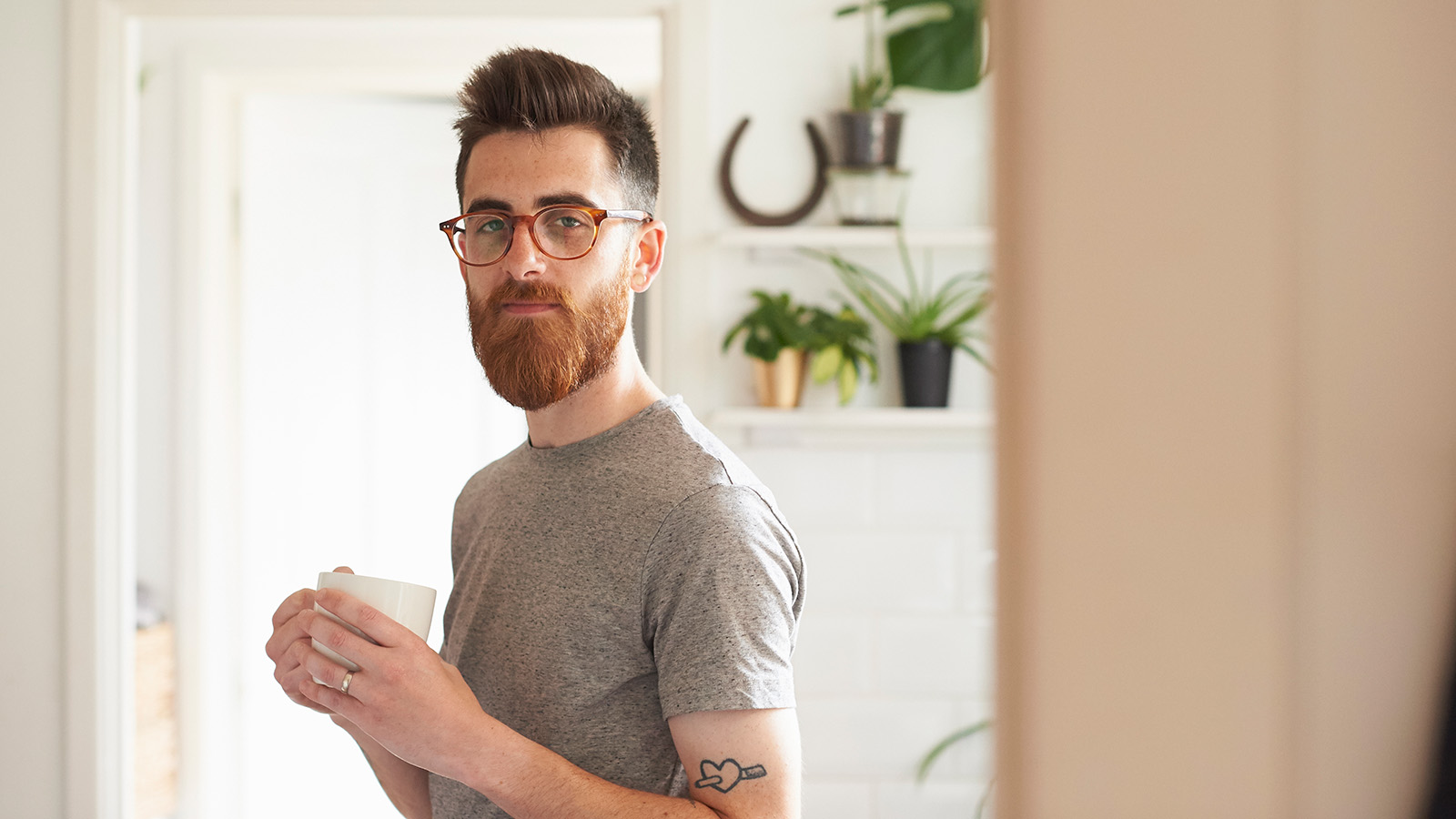 A man holds a cup in a bright room filled with plants, suggesting a moment of relaxation and contemplation.