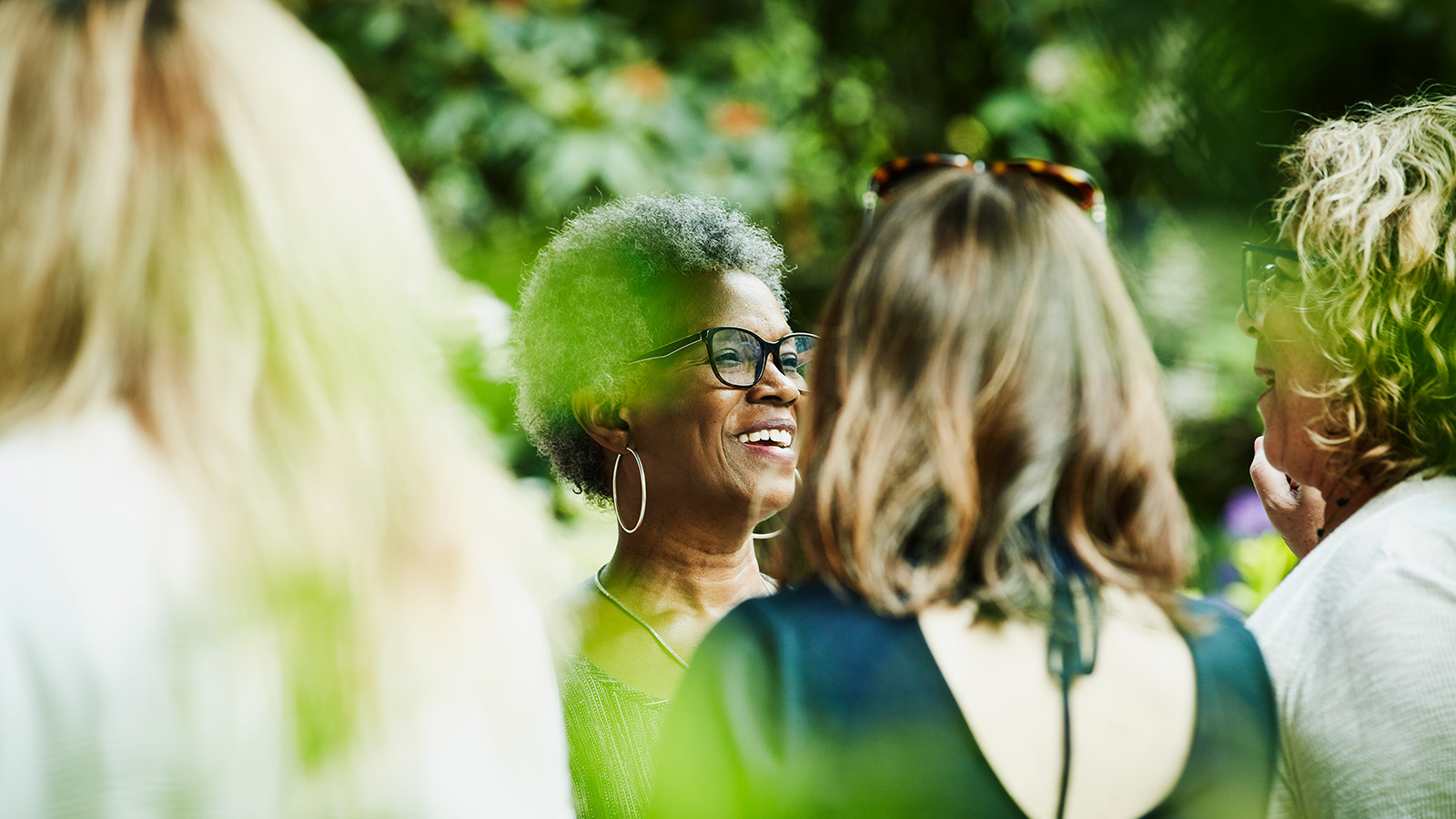 A woman with curly hair engages in conversation with a group of friends in a lush garden setting, surrounded by greenery.