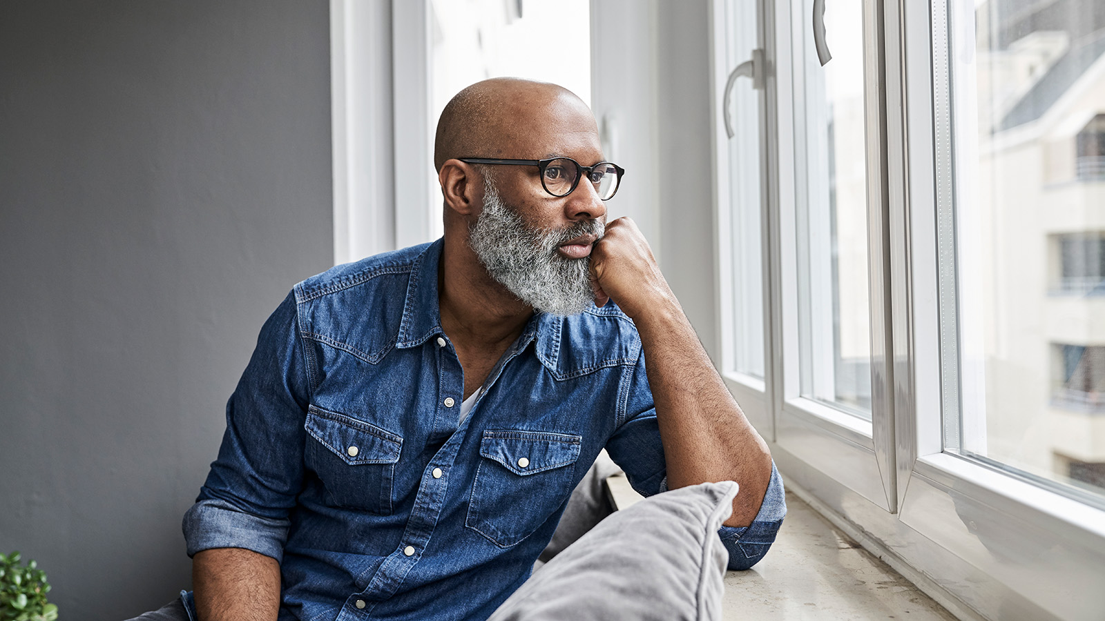 A man in a denim shirt gazes thoughtfully out a window, resting his chin on his hand in a cozy living space.