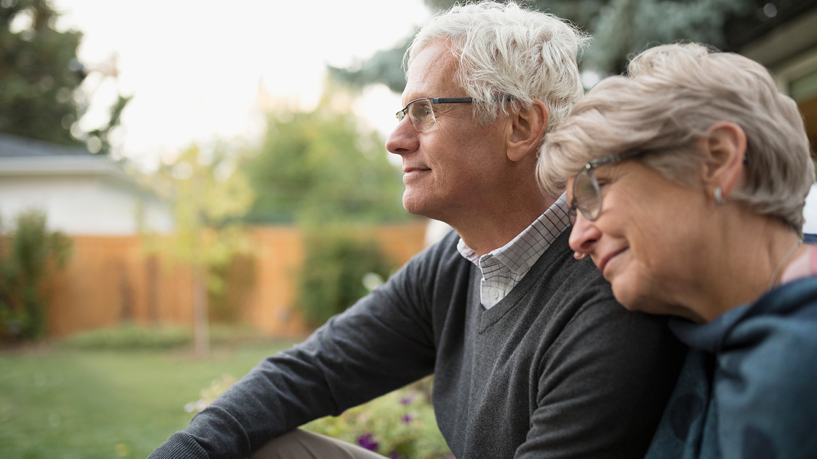 An older couple sits closely together on a bench in a garden, enjoying a peaceful moment outdoors in soft sunlight.