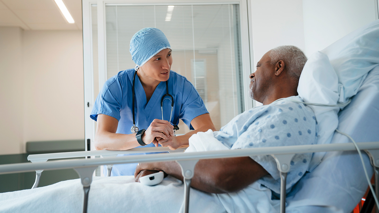 A nurse in scrubs holds a patient’s hand while discussing care in a hospital room with a patient in a hospital gown.