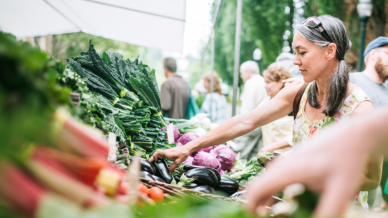 A woman is selecting vegetables at a market stall, surrounded by fresh produce and other shoppers in the background.
