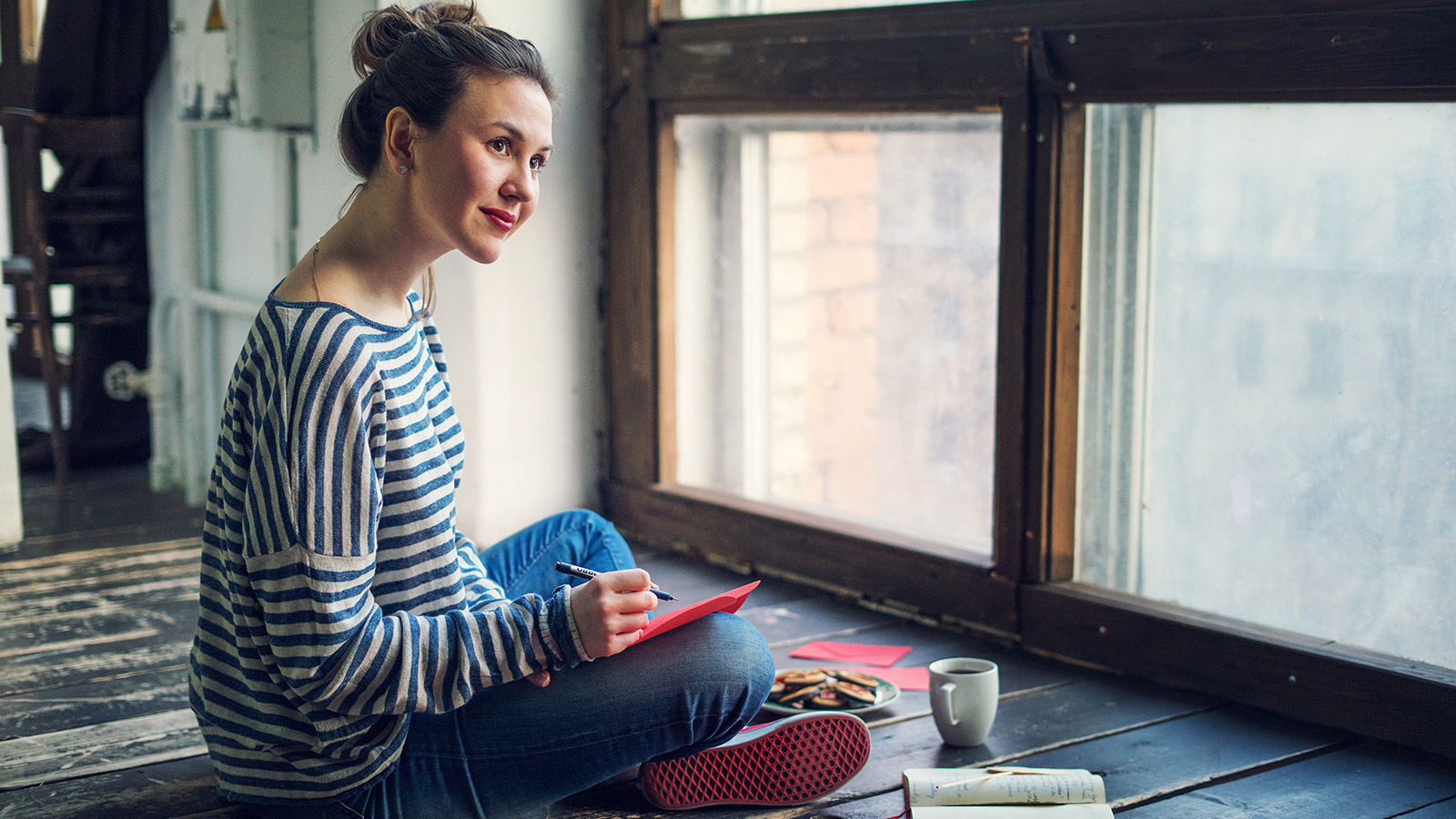 A woman sits on the floor, writing in a notebook, surrounded by snacks and a cup of coffee.