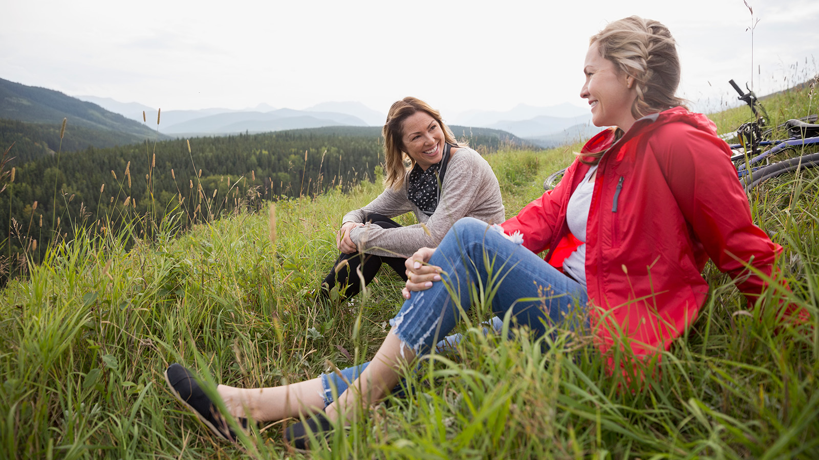 Two women are sitting in a grassy area, overlooking a scenic view of mountains and trees.