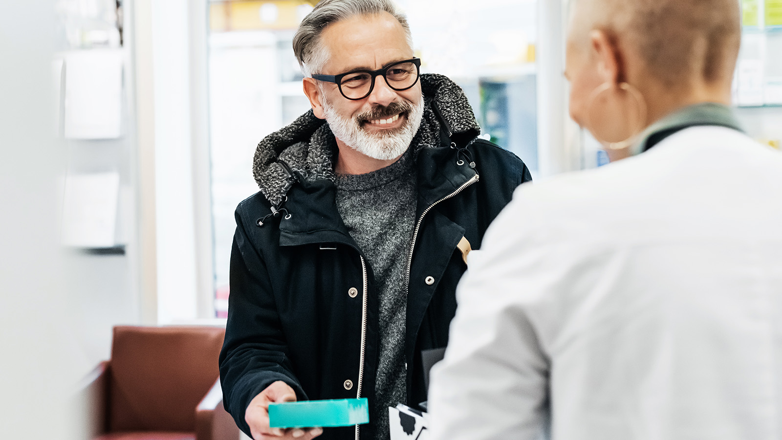 A man in a black coat receives a package from a pharmacist in a well-organized pharmacy setting.