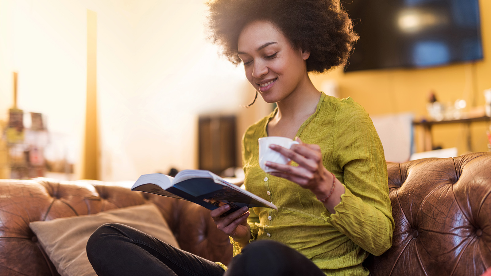 A woman with curly hair sits on a couch, reading a book and sipping from a cup in a warm, inviting room.