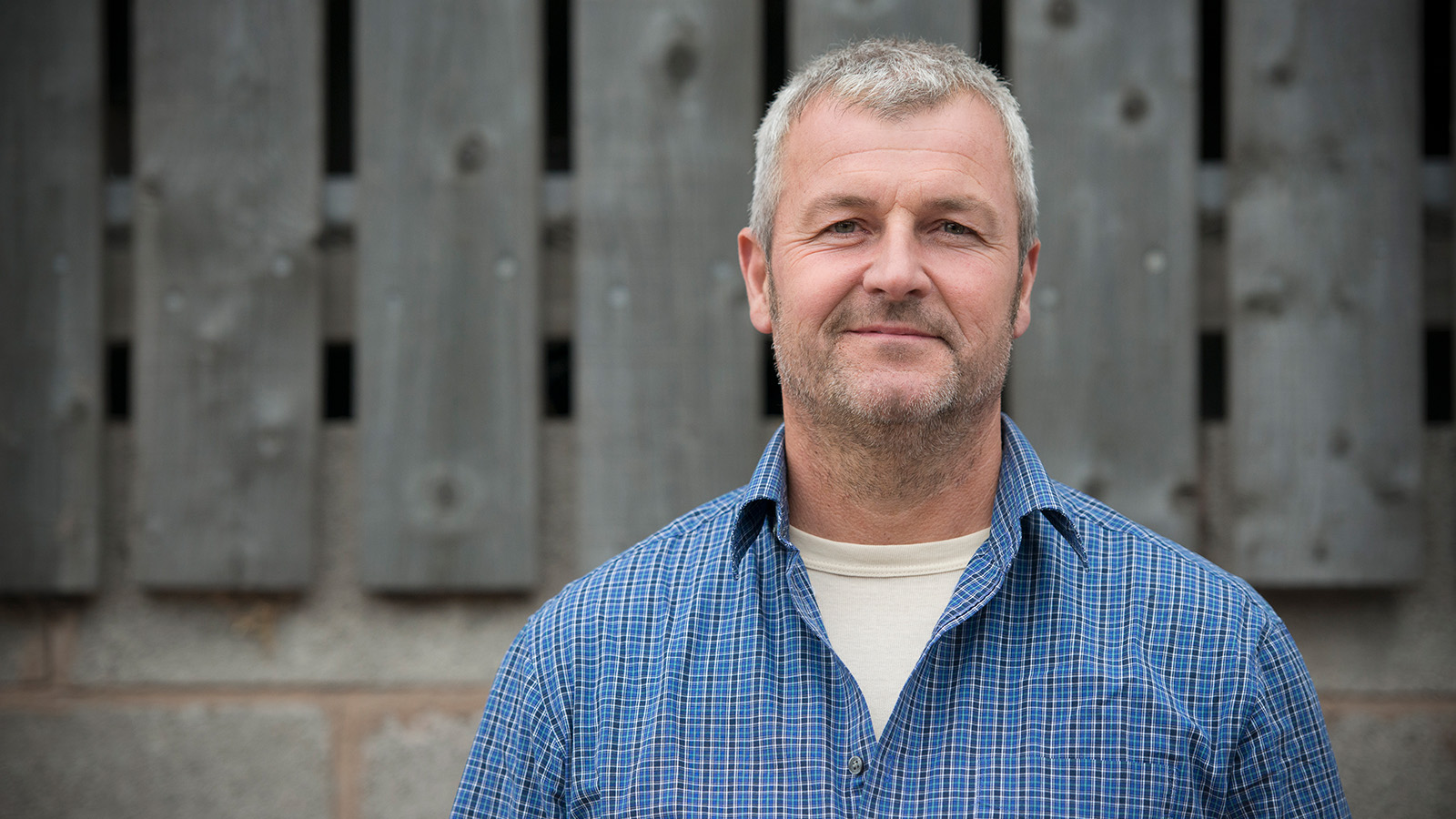 A man standing outdoors in front of a wooden fence, wearing a blue checkered shirt.