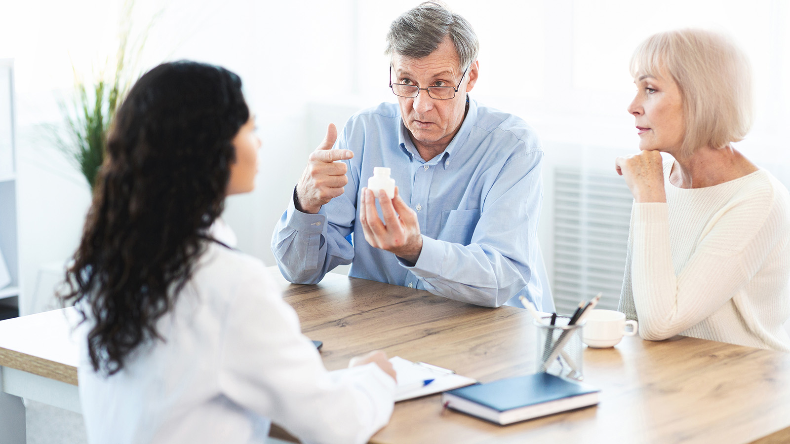 A doctor discussing medication with an elderly couple at a table, with a prescription bottle in the man’s hand.