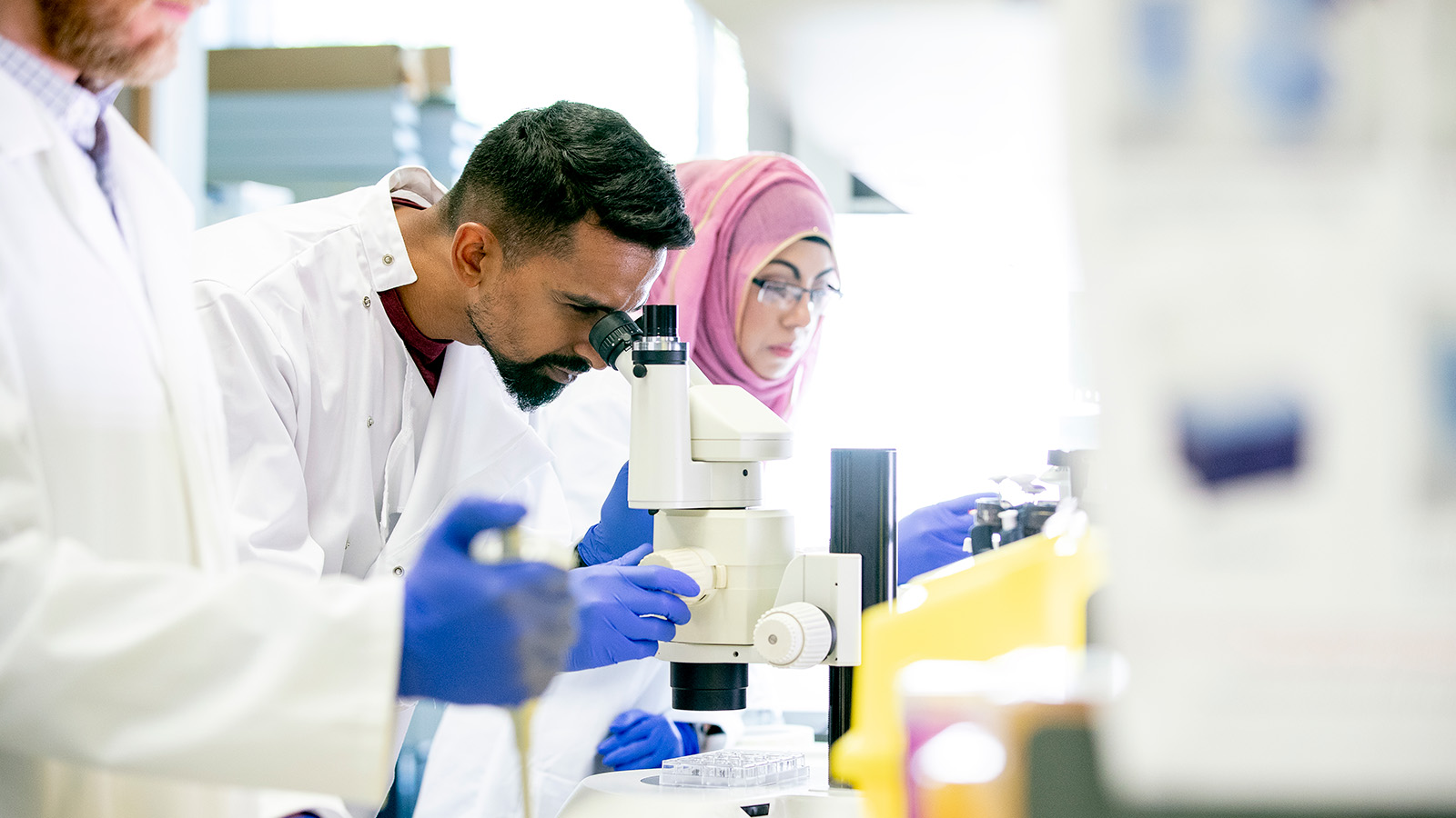 A group of researchers in lab coats working together at a laboratory table with a microscope.