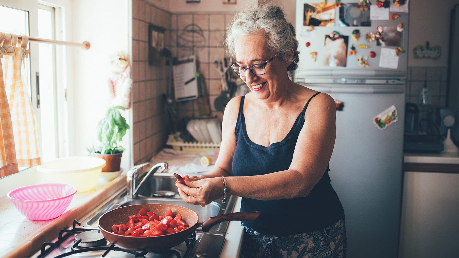 A woman cooking in a kitchen, preparing vegetables in a frying pan, with various kitchen items visible.