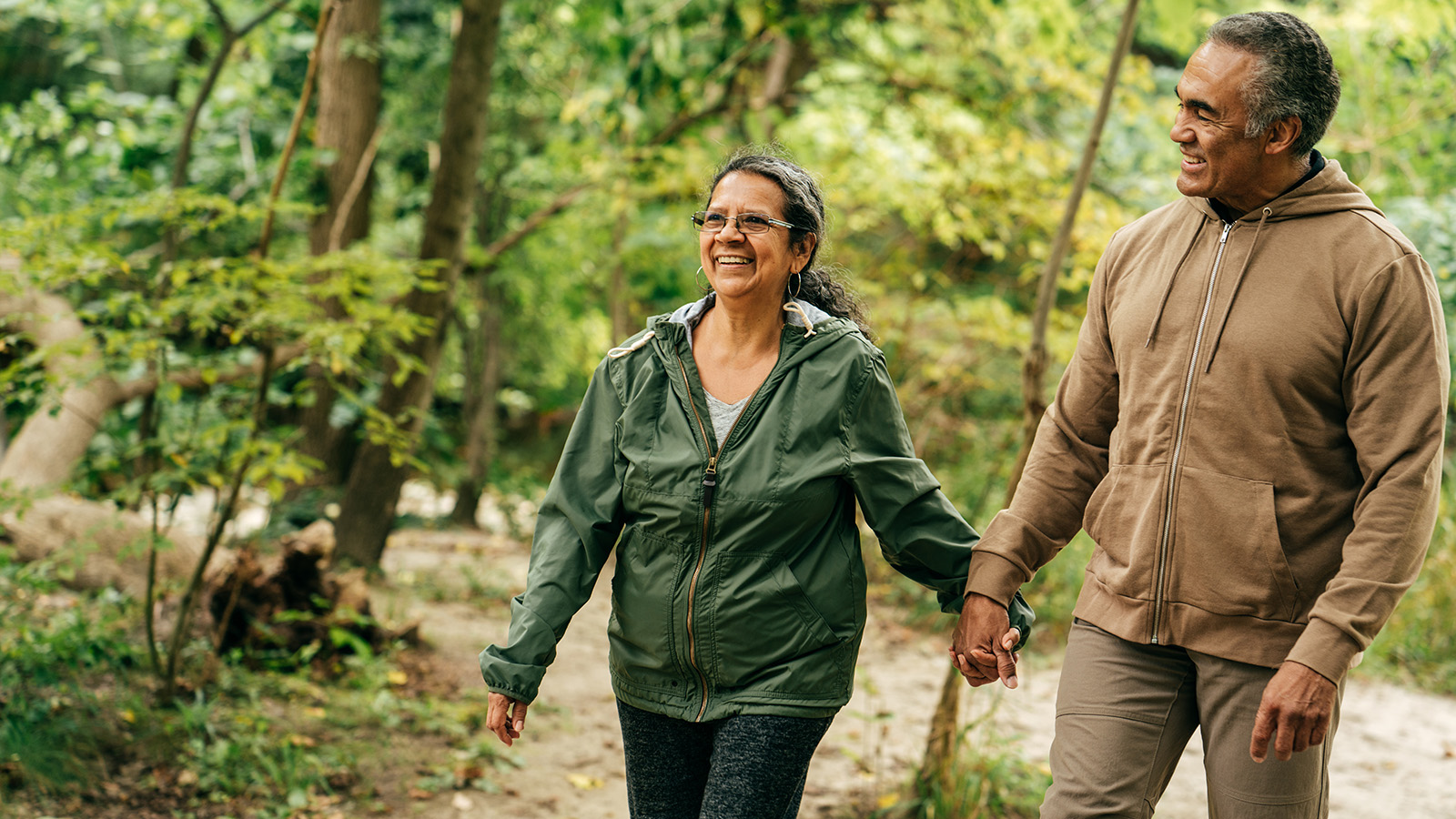 A couple walking hand-in-hand through a lush green forest, both wearing casual outdoor clothing.