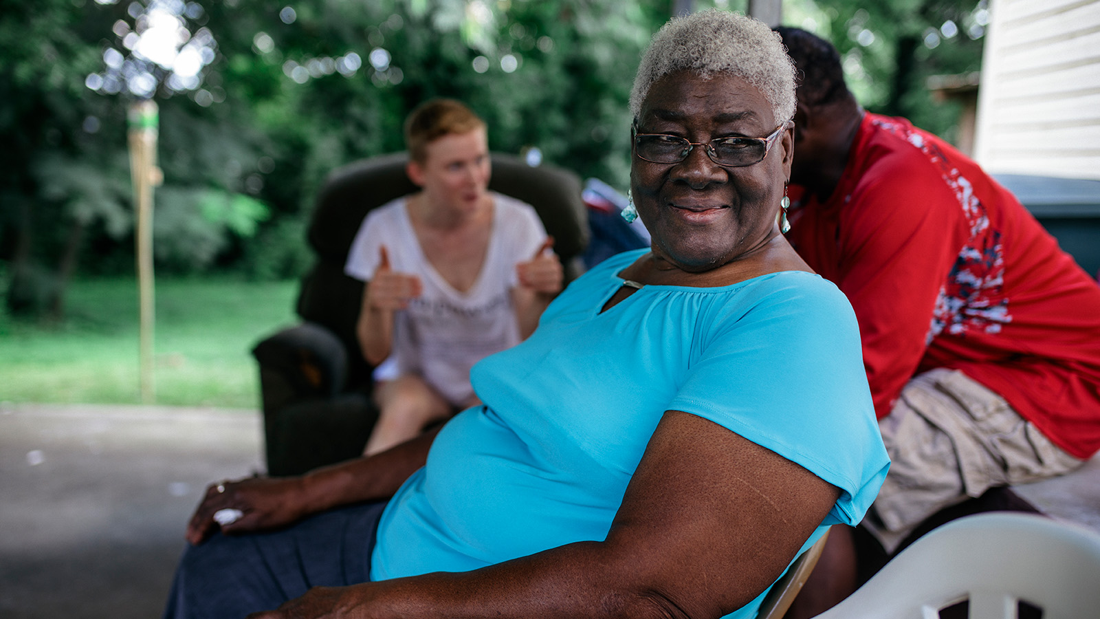 A woman in a blue shirt sitting on a porch, surrounded by friends, with greenery in the background.