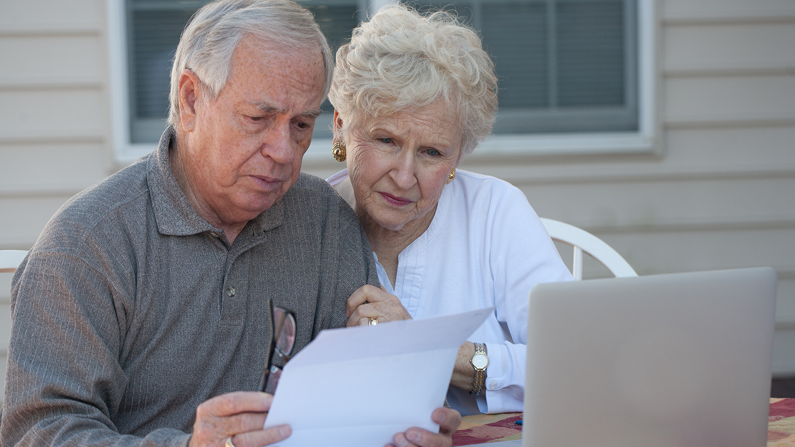 An elderly couple sitting together outdoors, looking at a letter while a laptop is open on the table.