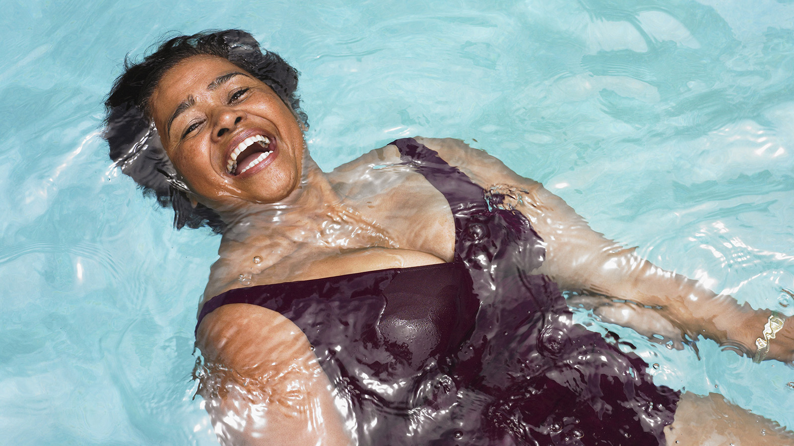 A woman floating in a swimming pool, water rippling around her, wearing a dark swimsuit.
