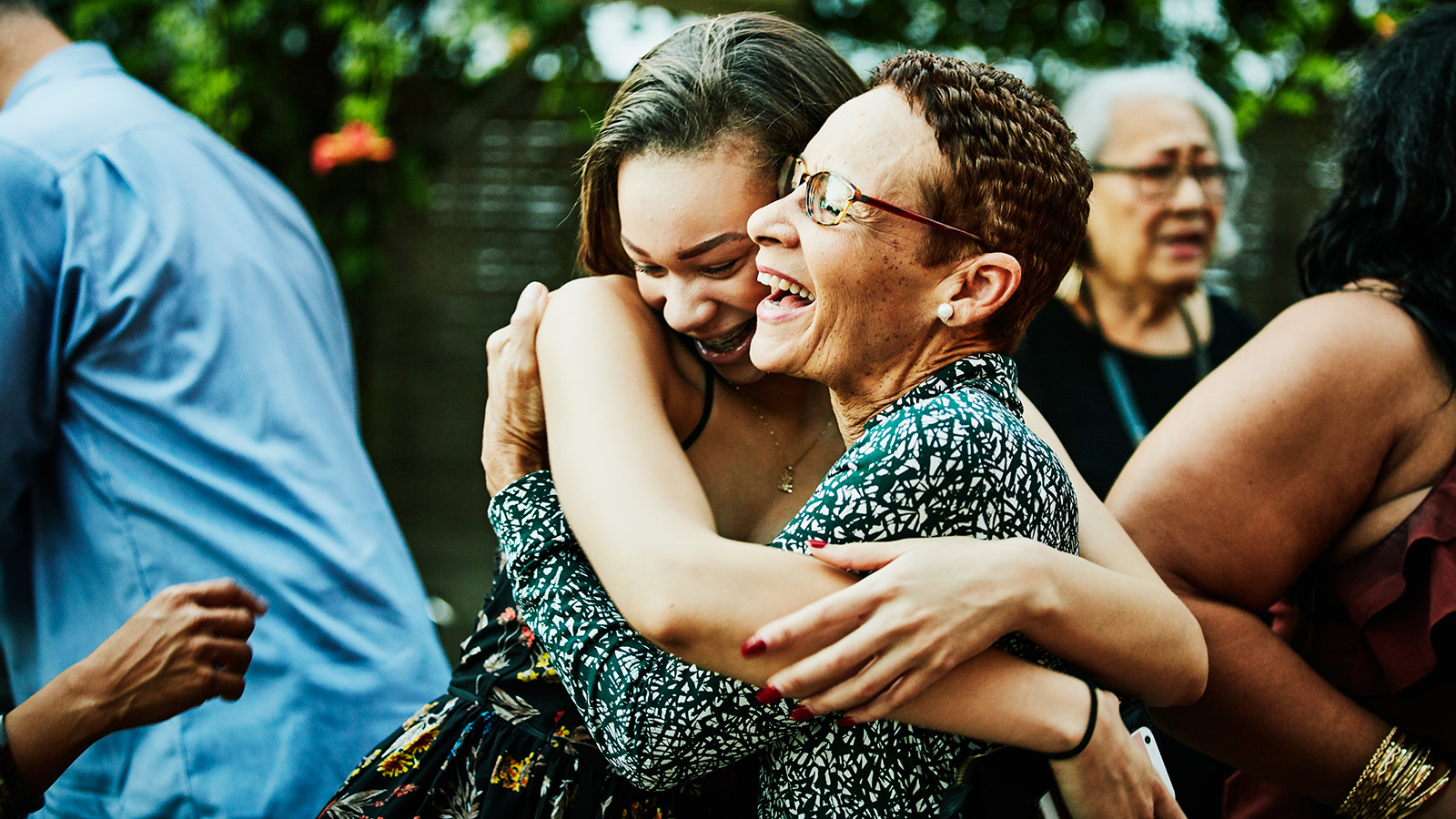 Two women embracing each other outdoors, with trees and houses visible in the background.
