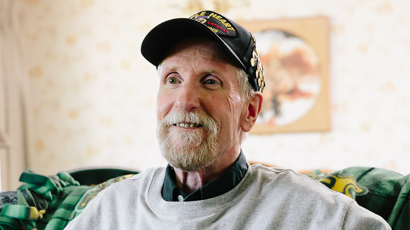 An elderly man sits on a couch, wearing a cap and a gray sweatshirt, surrounded by cozy blankets and decor.