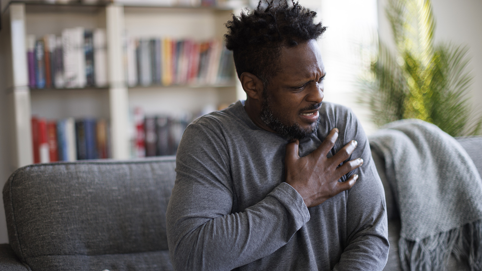 A man sits on a couch, holding his chest, appearing distressed in a cozy living room.