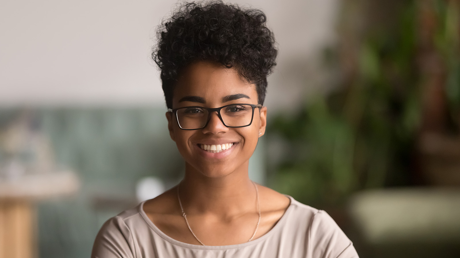 A woman sits calmly, looking directly at the camera, with a soft background of greenery.
